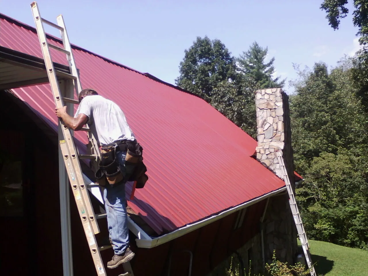 Roofer on a ladder beside a red metal roof and stone chimney — Soffit & Fascia Repair by Roofing Brothers in Thompsonville